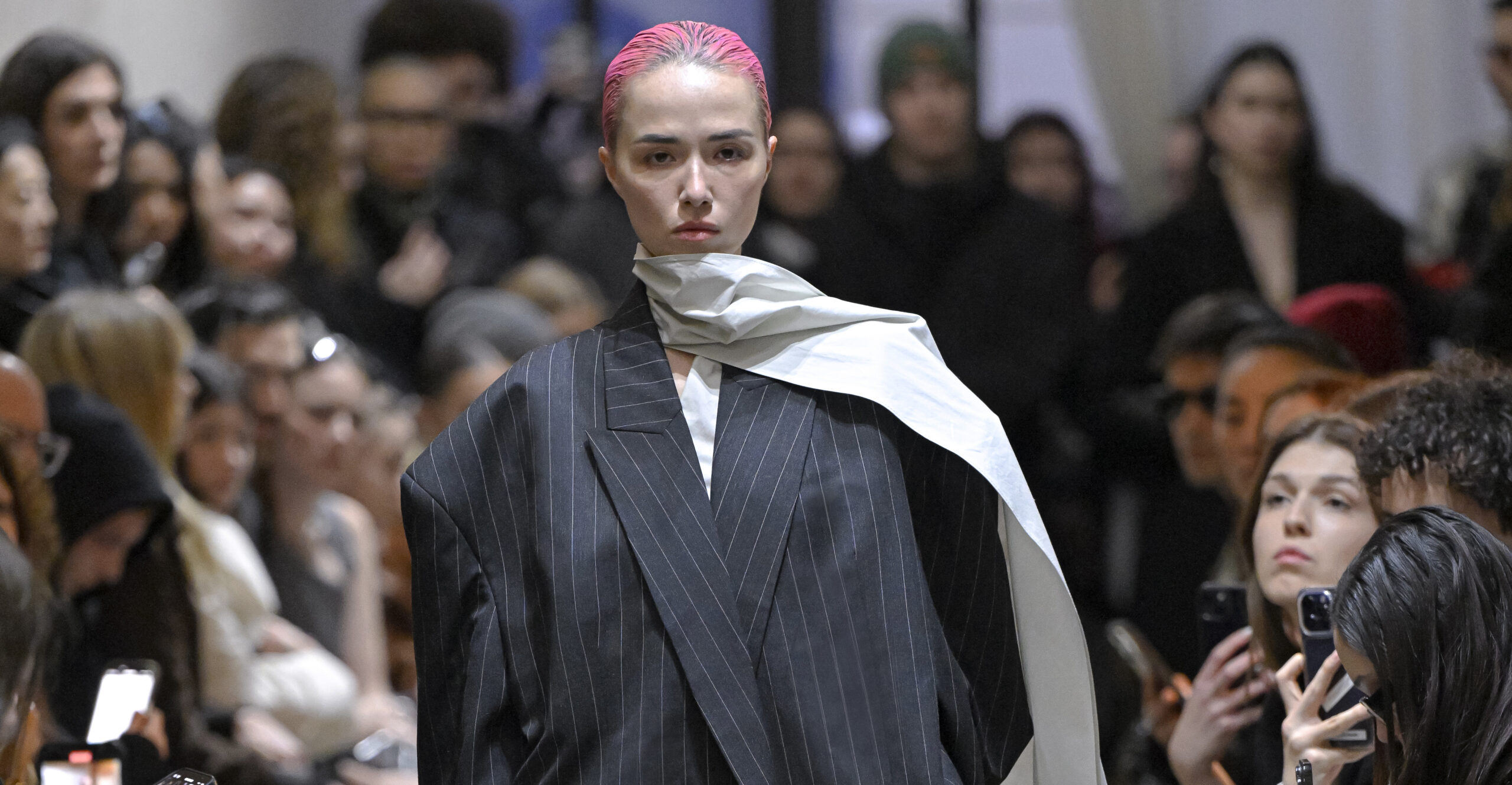 A model exhibits a bold pinstripe blazer with a draped cream scarf from Vautrait's Fall/Winter 2024 collection on the runway, complemented by a stark pink hair color, encapsulating the brand's avant-garde approach to sustainable fashion.
