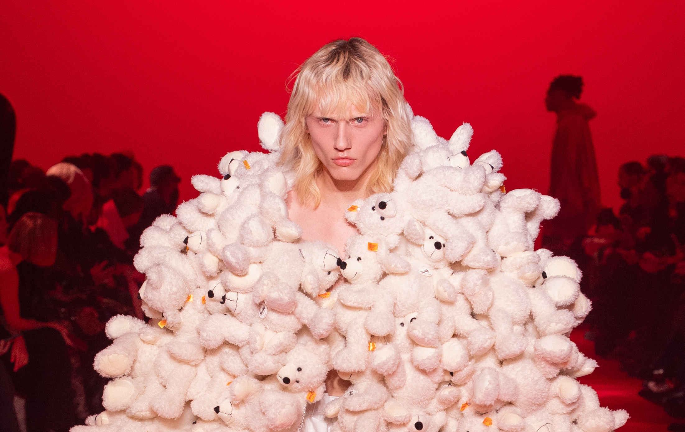 A model on the runway is enveloped in an extravagant dress made of plush teddy bears, against a vivid red backdrop.