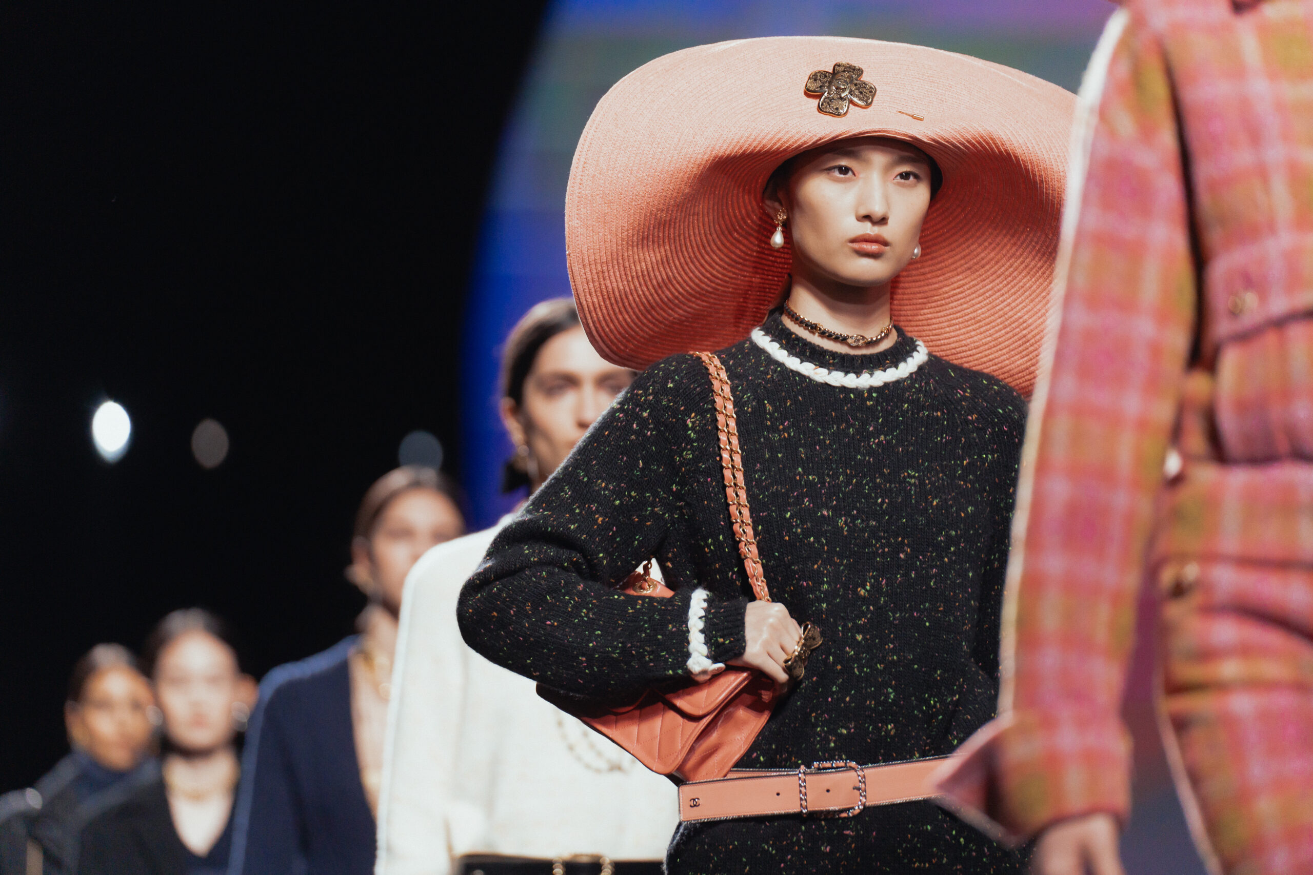 A model at the CHANEL Fall/Winter 2024/25 show is featured wearing a black speckled tweed dress with white collar and cuffs, accessorized with a large peach-colored hat, pearl necklace, and a matching crossbody bag.