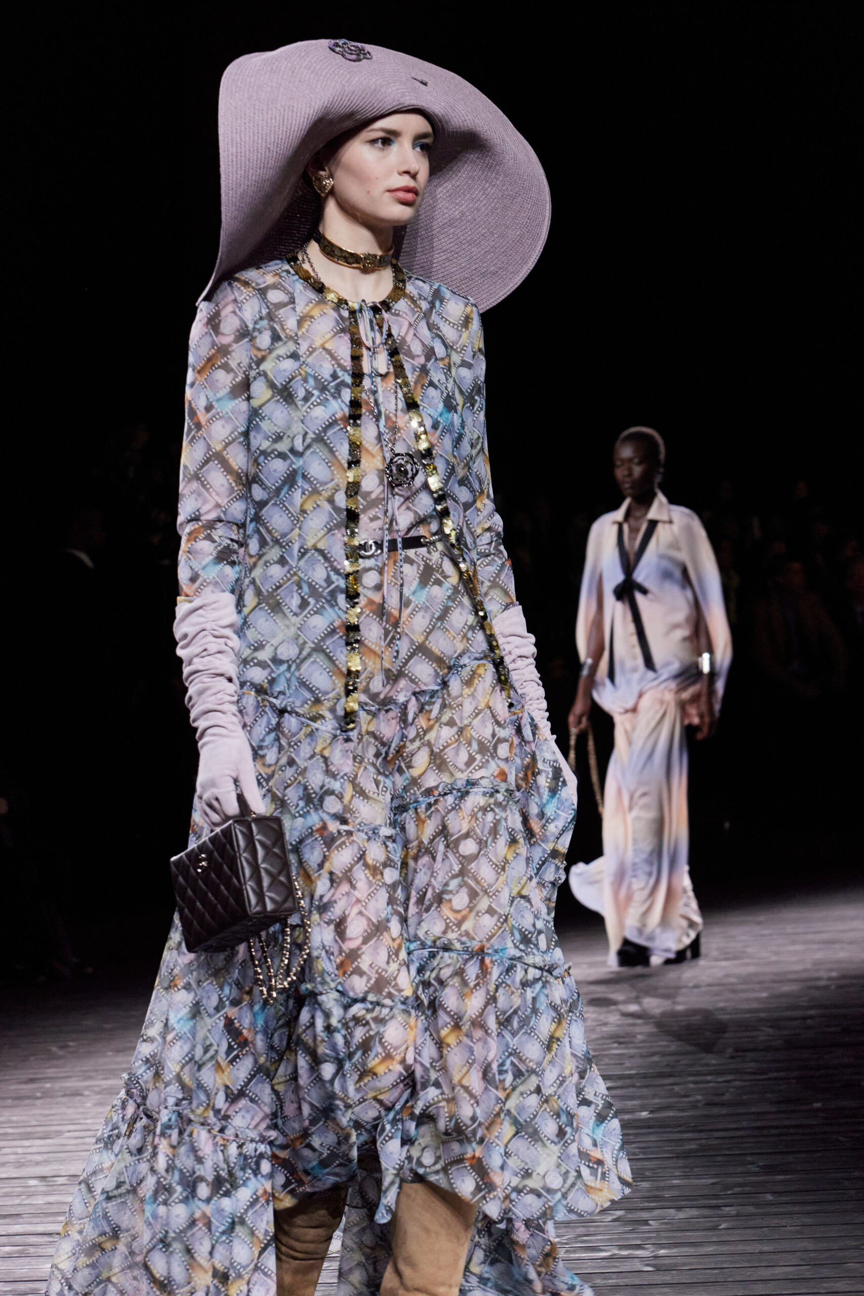 A model walks the runway at the CHANEL Fall/Winter 2024/25 show in a pastel patterned dress with film motif, paired with a wide-brimmed hat, accessorized with layered necklaces and a quilted handbag.