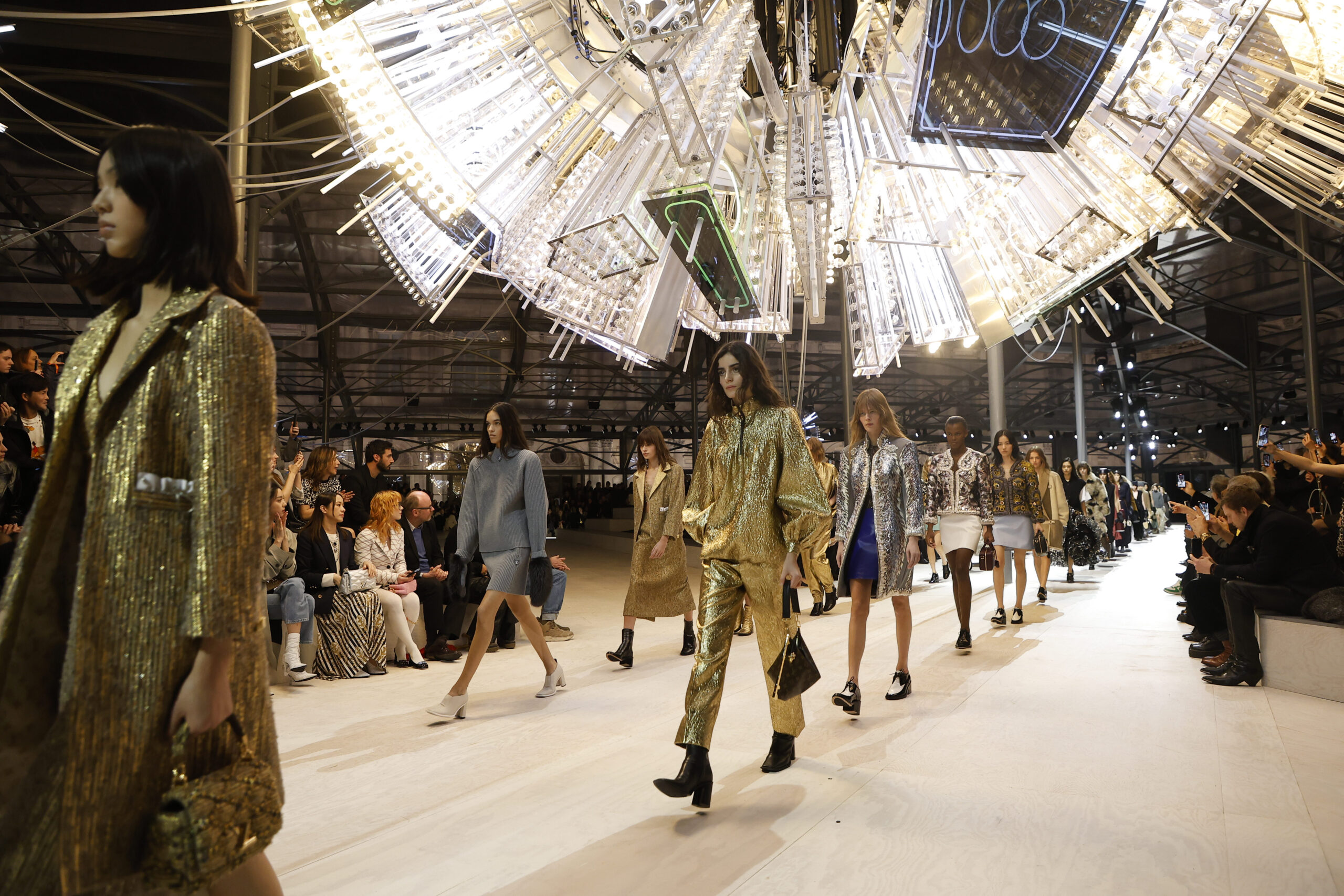 Models walk down the runway under dazzling chandeliers, showcasing a variety of Louis Vuitton ensembles in a spectacle of light and fashion.