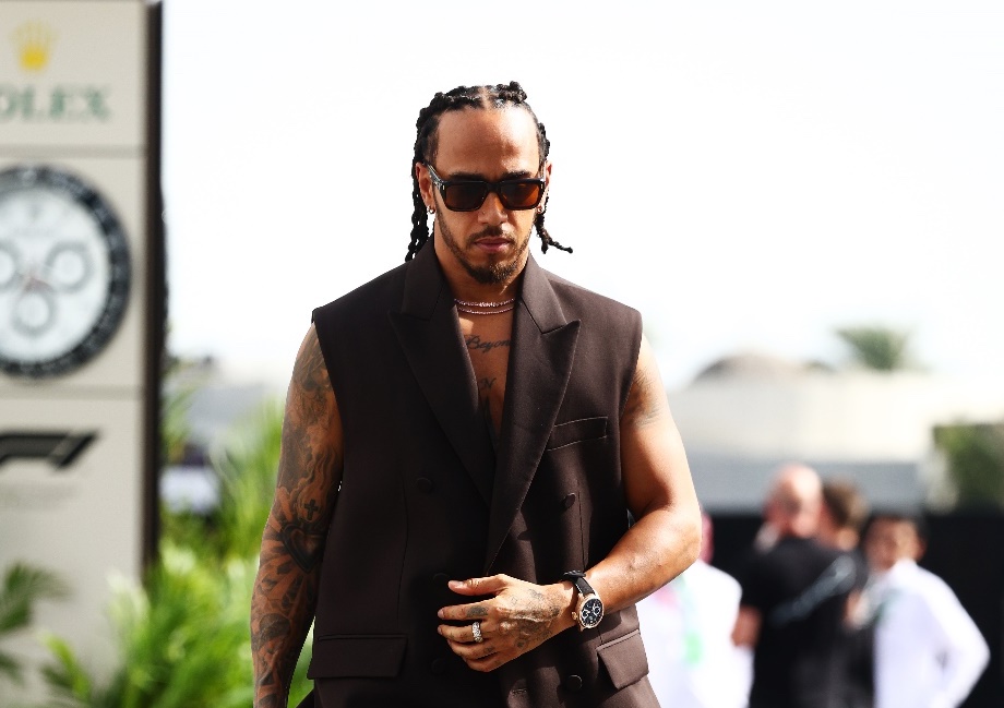 Lewis Hamilton walks confidently in a sleeveless, chocolate brown wool coat and matching trousers from Gucci's Men’s Fall Winter 2024 collection. He pairs his outfit with black leather Gucci mocassins featuring the Horsebit detail, as he strides through the paddock before the F1 Grand Prix of Saudi Arabia.