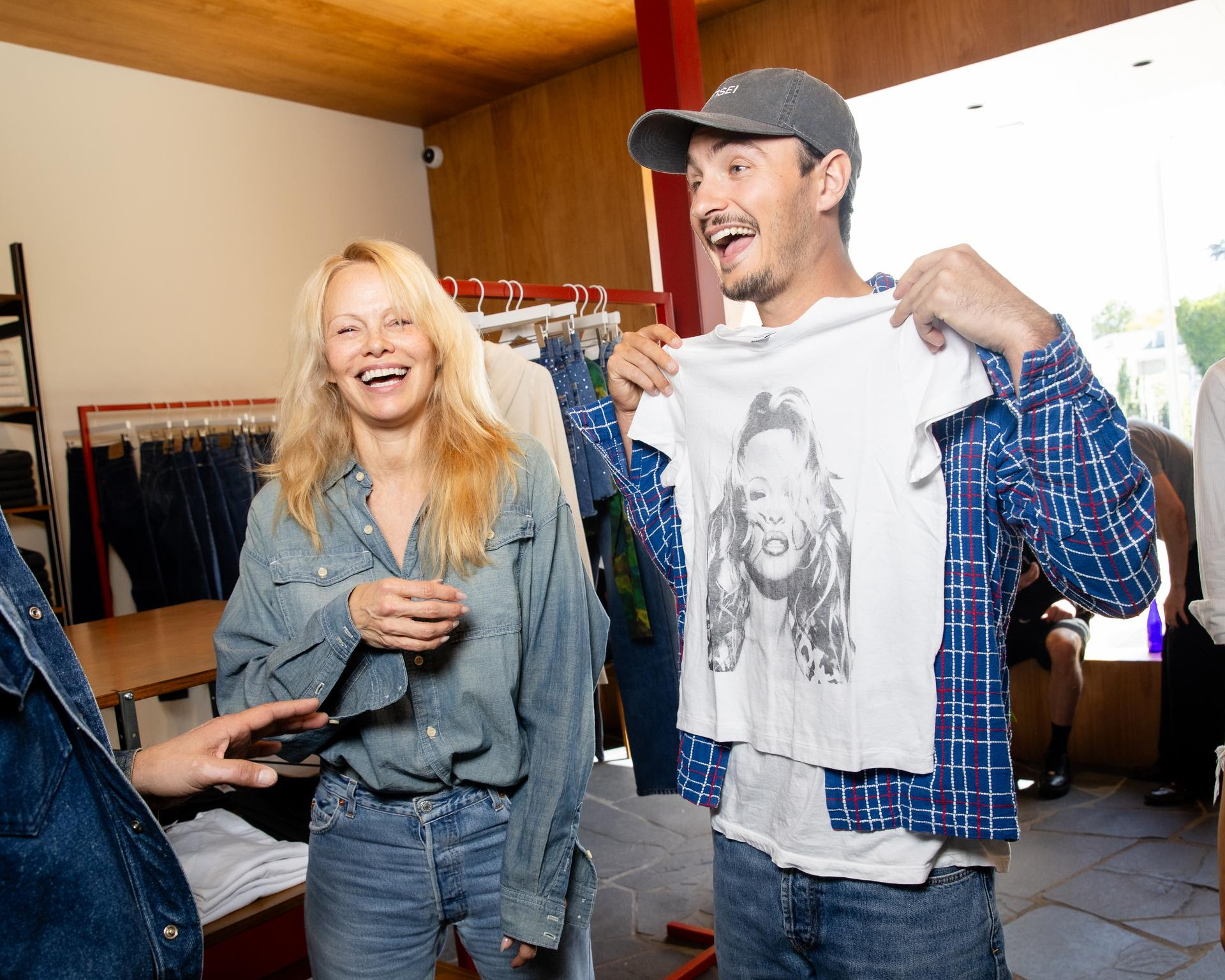 Pamela Anderson laughs joyfully beside her son Brandon Thomas Lee, who holds up a t-shirt featuring her iconic image, at the RE/DONE x Pam event.