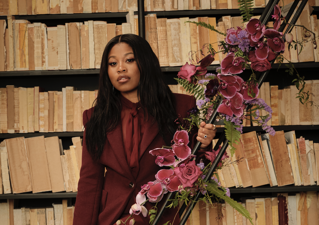 Dominique Fishback stands confidently in a maroon Sergio Hudson suit, holding a floral arrangement, against a library backdrop at the Creative Collective NYC event.